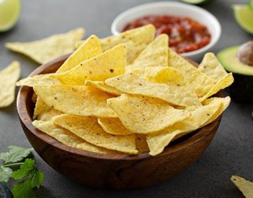 Wooden bowl filled with crispy yellow tortilla chips with salsa and avocado in the background